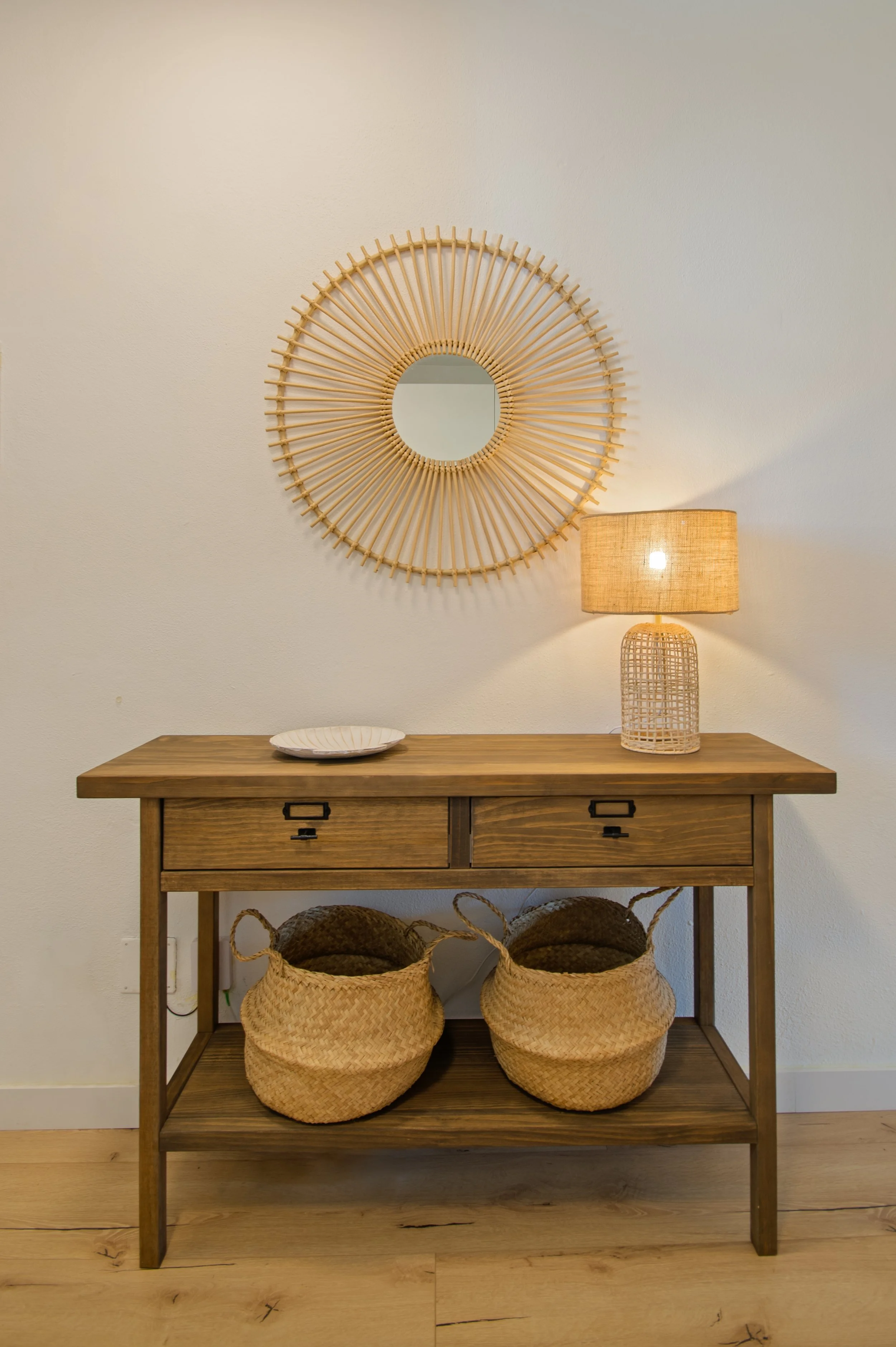 Mesa de madera con tres cajones, dos cestas de mimbre en la parte inferior, un plato blanco sobre la mesa, un espejo de ratán en la pared y una lámpara de mimbre con luz encendida.