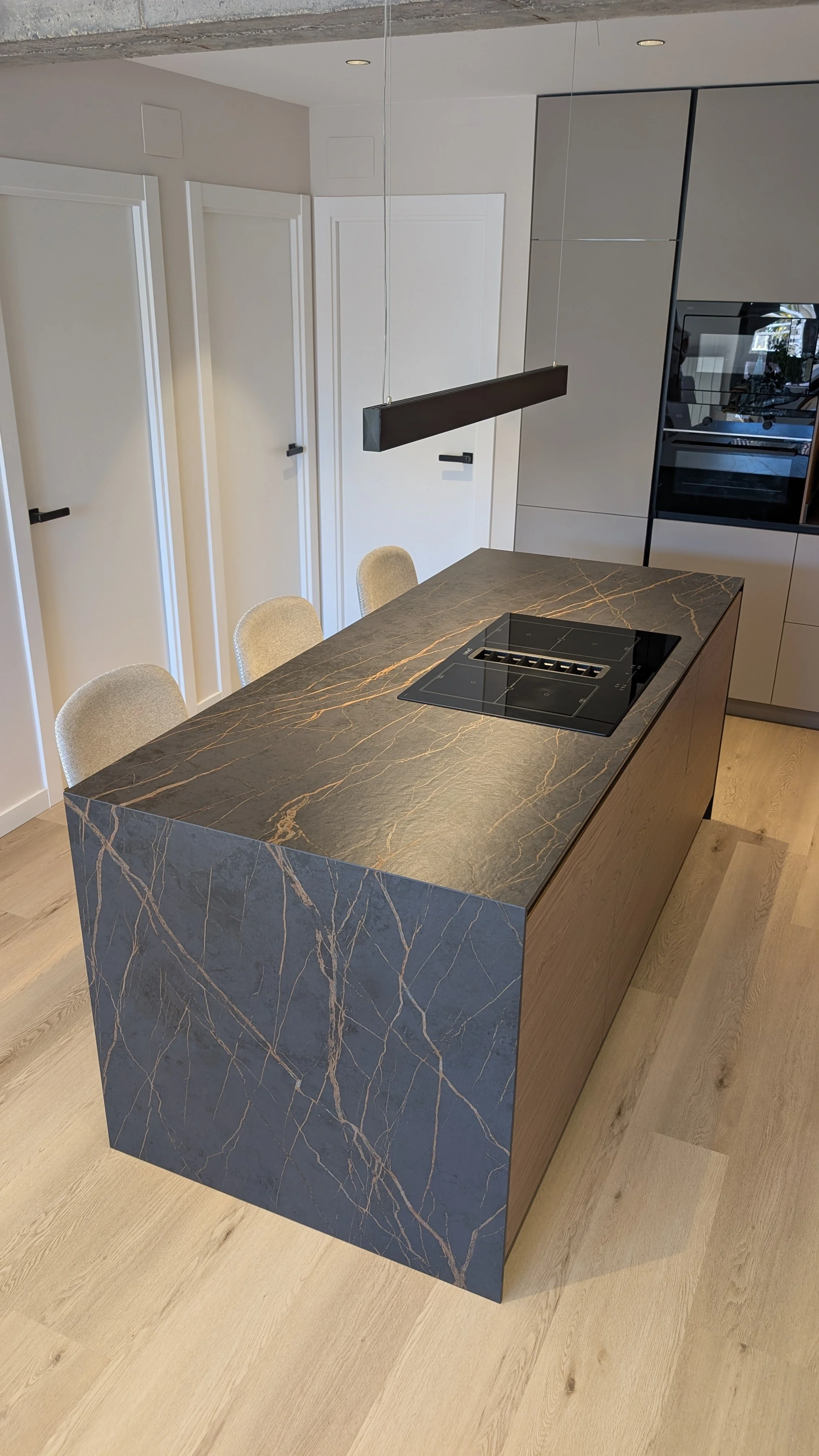 Cocina moderna con isla de mármol oscuro con vetas doradas, tres sillas beige, armarios grises y un electrodoméstico empotrado en la pared.