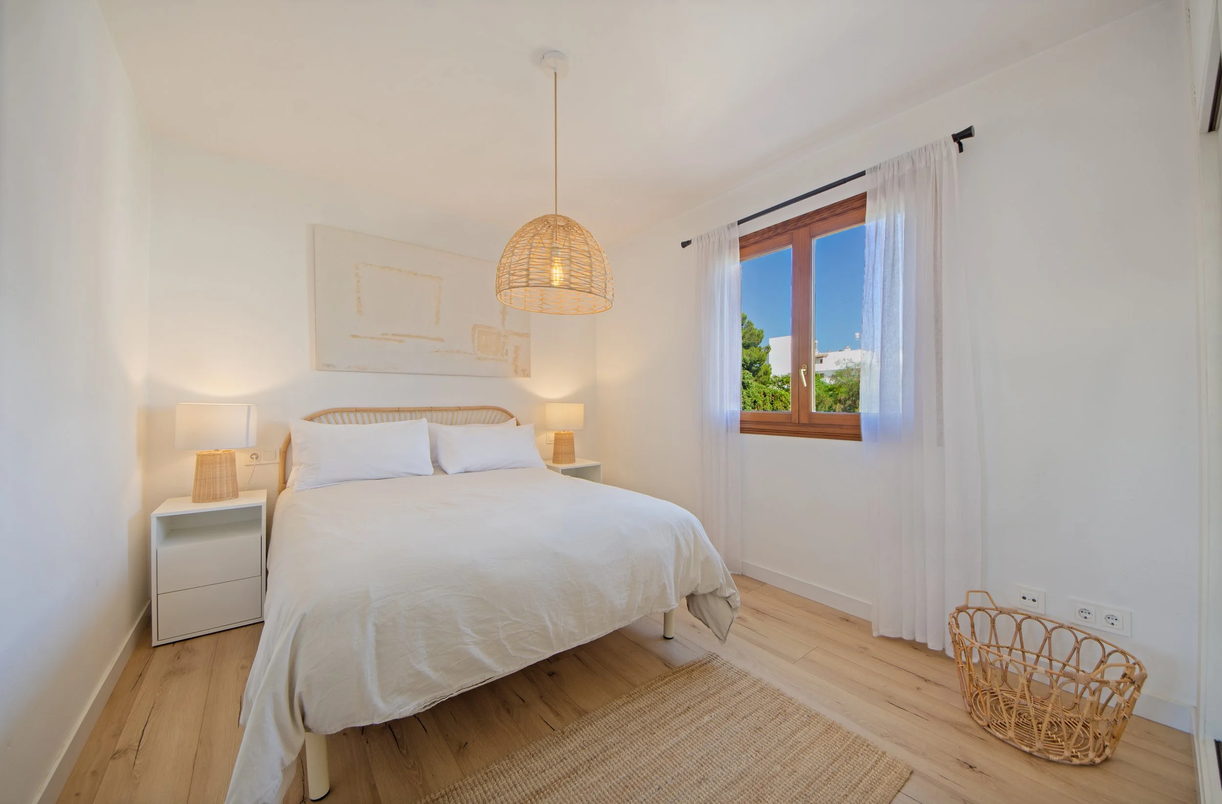 interioristavitoriagazteizHabitación con cama blanca, dos mesas de noche con lámparas, ventana con cortinas blancas, decoración minimalista y salida a un espacio con árboles verdes.