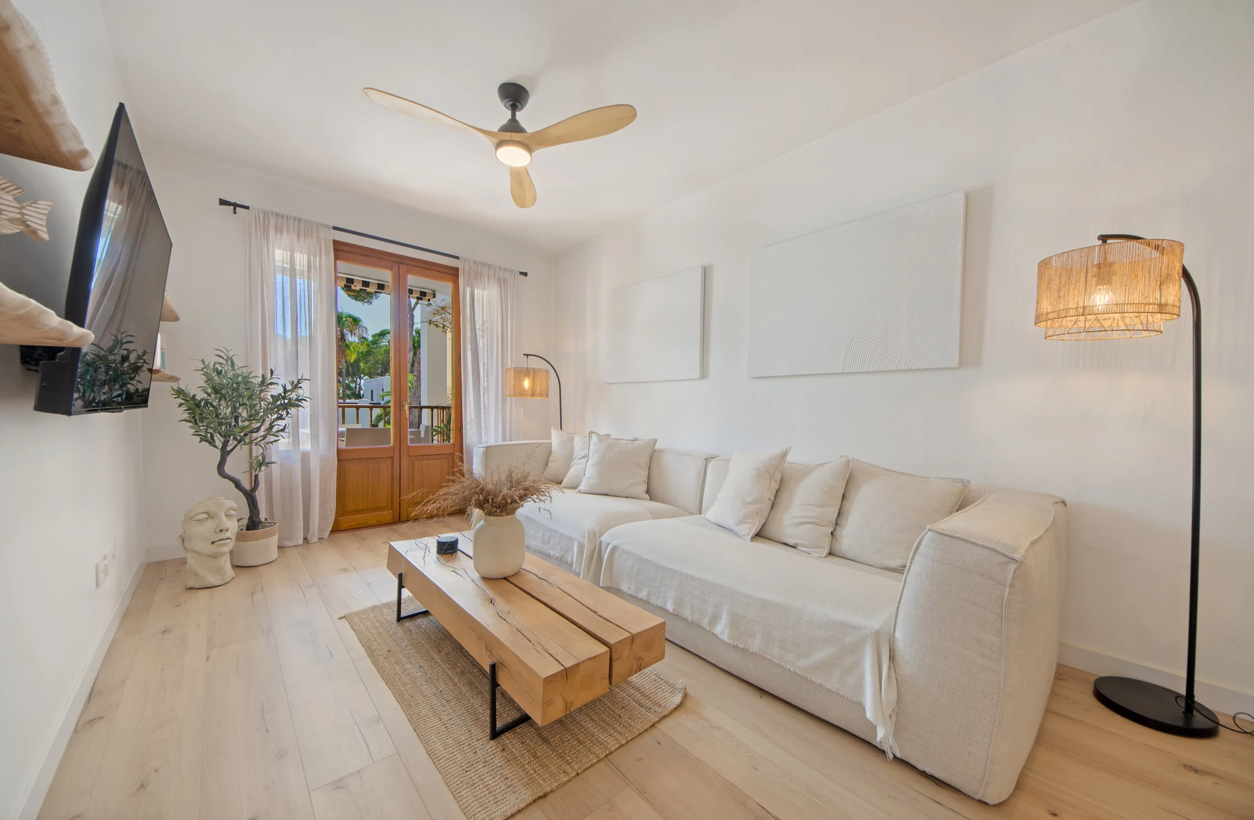 Sala de estar con sofá grande de color gris claro, mesa de madera rectangular, lámpara de piso con pantalla de fibra natural, cuadros blancos en la pared, ventanas grandes con cortinas blancas, puerta de madera con vidrio, planta y decoraciones en la