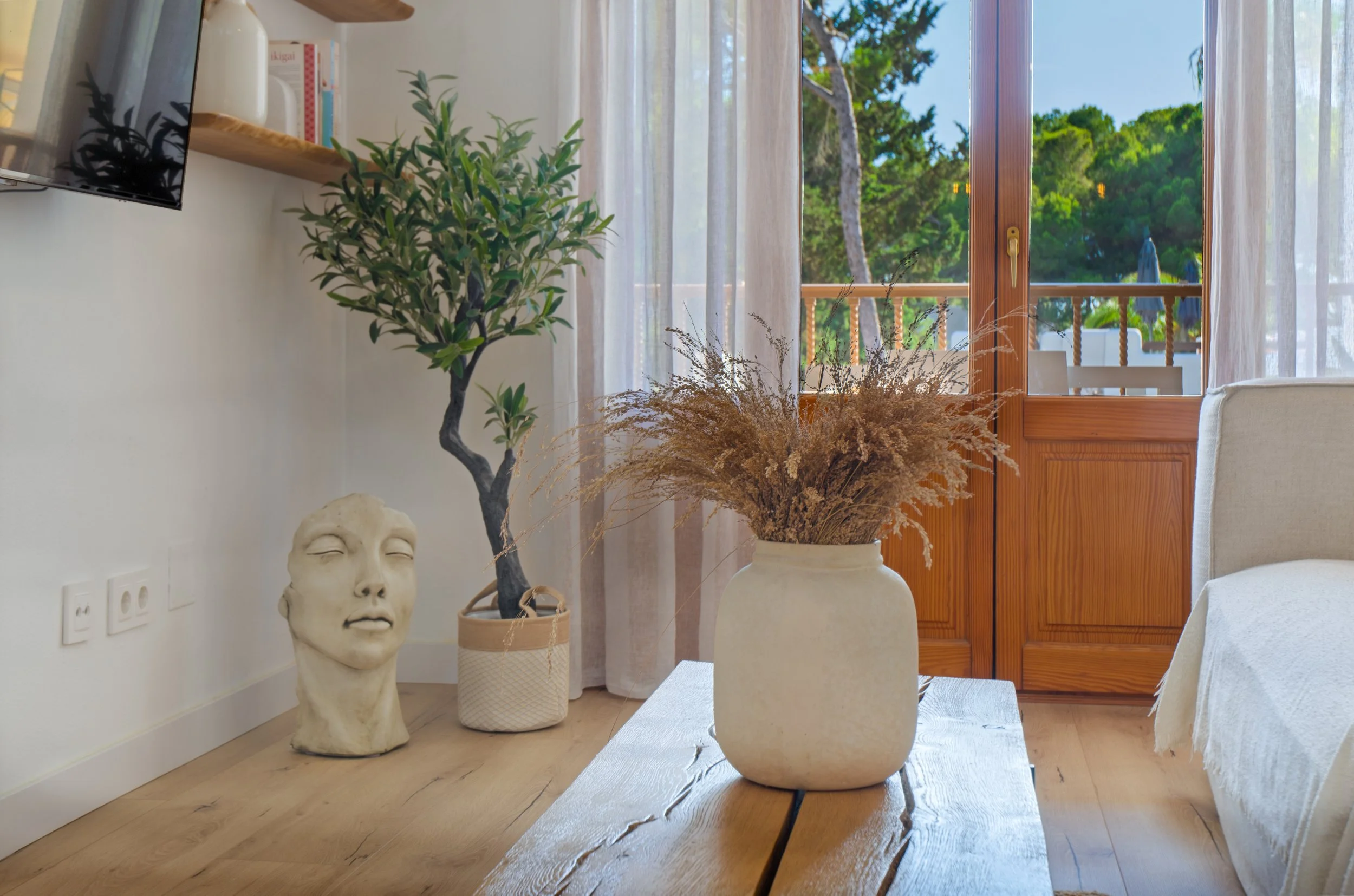 decoraciónseoaoneinteriorismo Interior de una sala con varias plantas en macetas, una escultura de rostro humano, un televisor en la pared, cortinas blancas, una puerta de madera que da a un balcón con vista a árboles verdes y sillones.