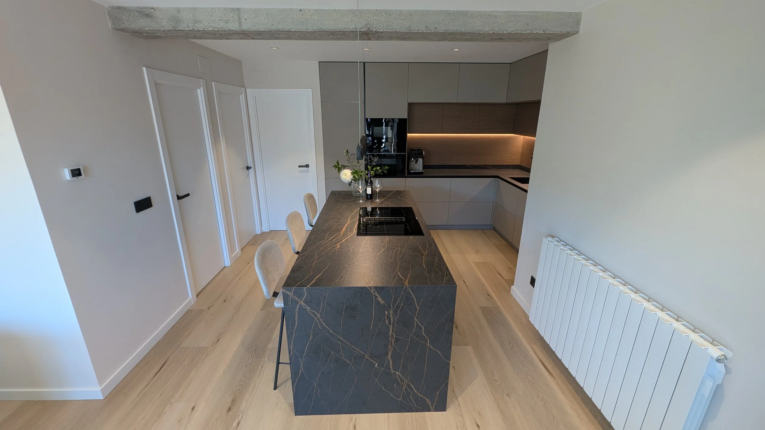 Cocina moderna con gabinetes grises y una isla de mármol negro con vetas doradas, paredes blancas y suelo de madera clara diseñado por seoane interiorismo en vitoria-gazteiz.