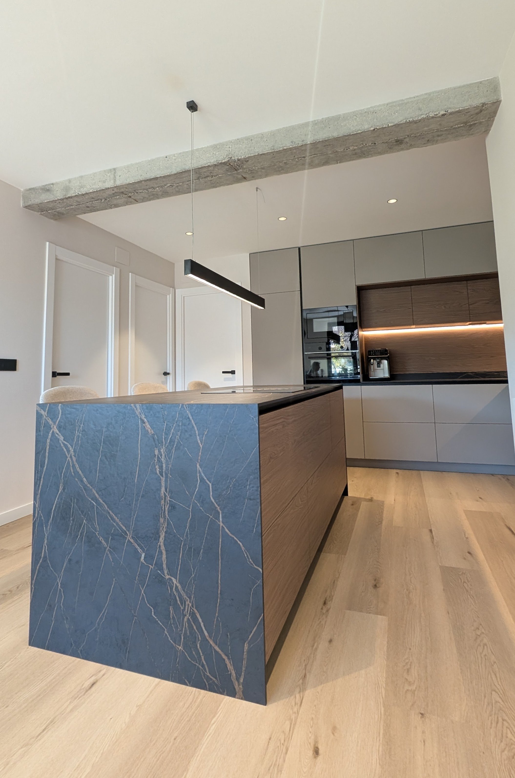 Cocina moderna con isla de mármol azul y gabinetes de madera y gris, con electrodomésticos empotrados y una lámpara de diseño recto colgando del techo fabricada y diseñada por Seoane Interiorismo vitoria-gazteiz