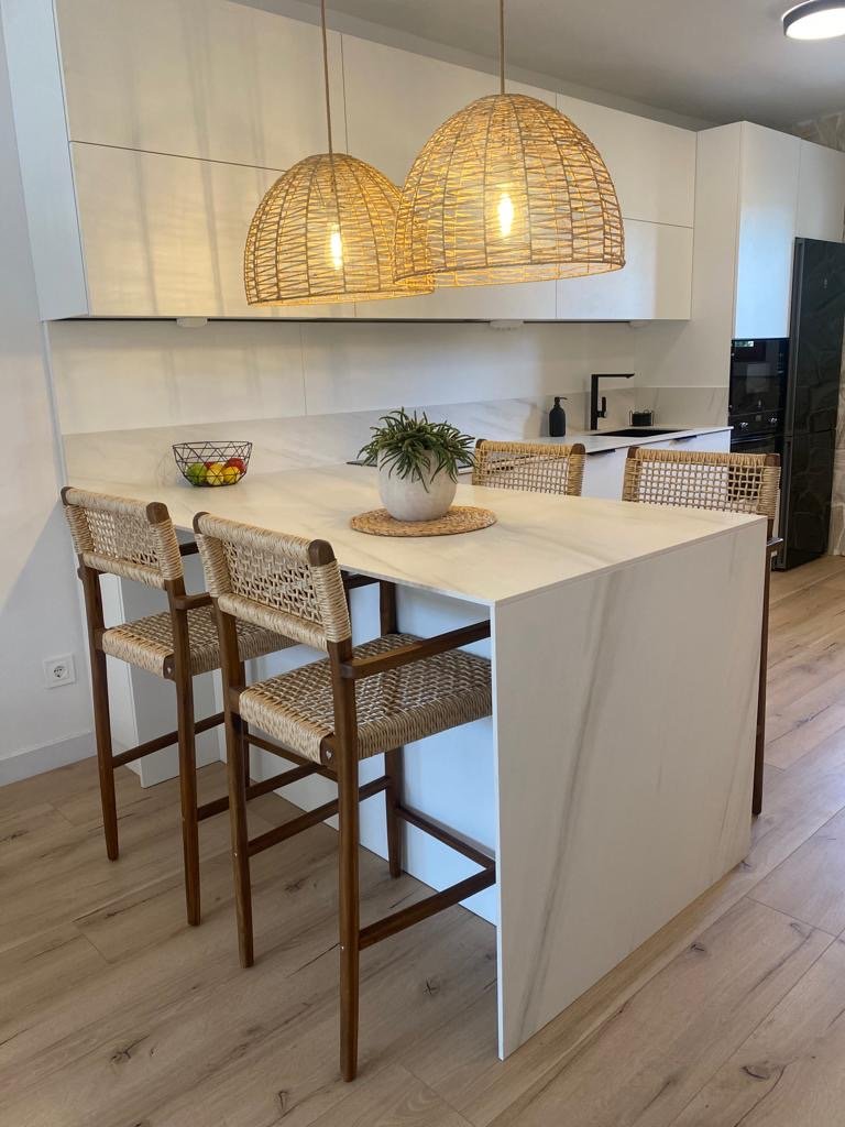 Una cocina moderna con una mesa de isla de mármol blanco, cuatro sillas altas de madera con asientos de mimbre, colgantes de mimbre y un potted de planta en el centro de la mesa diseñada por seone interiorismo en mallorca. 