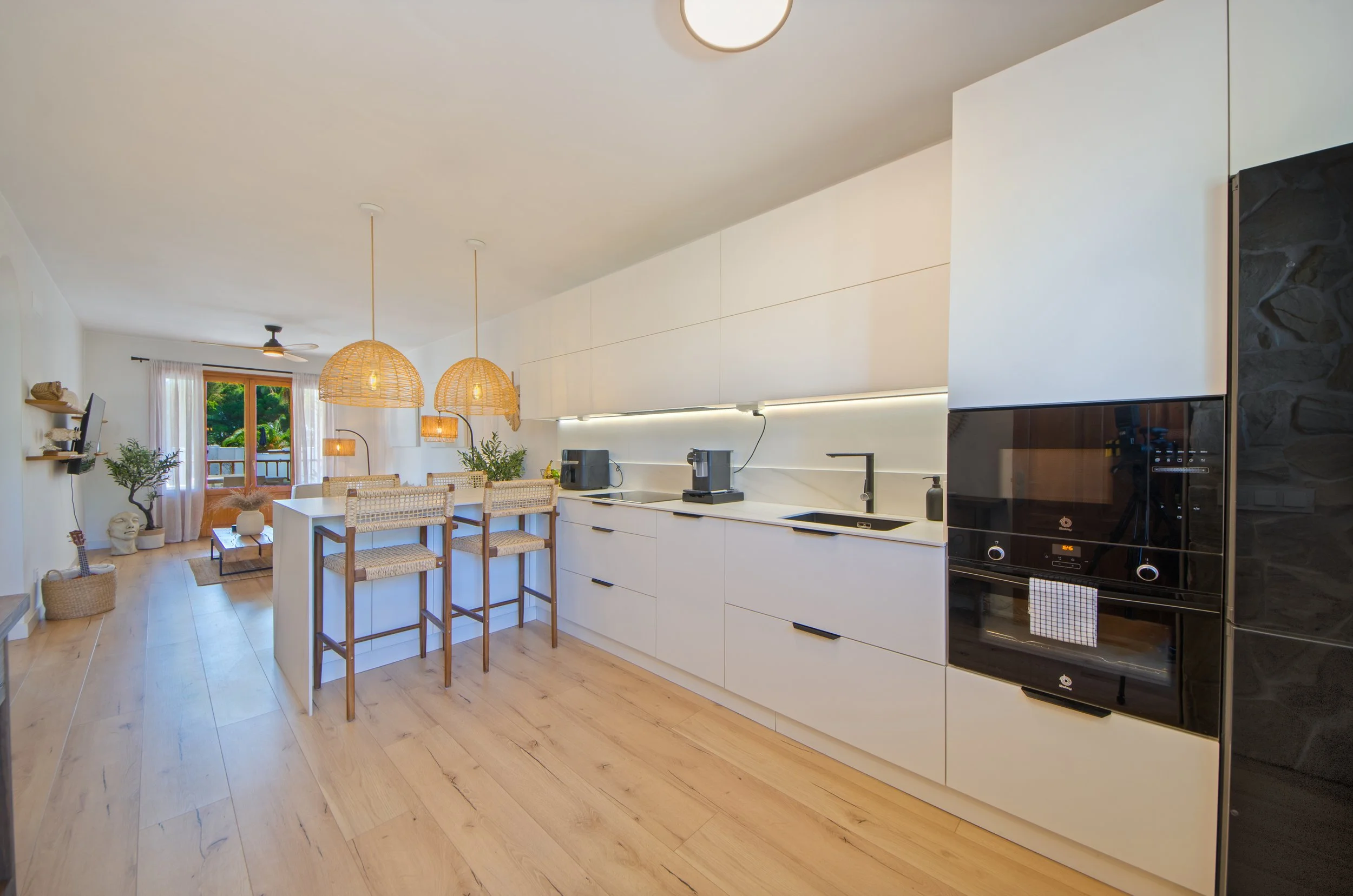 Cocina moderna con isla central, gabinetes blancos, electrodomésticos negros, mesas de madera, sillas de mimbre, lámparas de mimbre, plantas y vista a la terraza exterior.