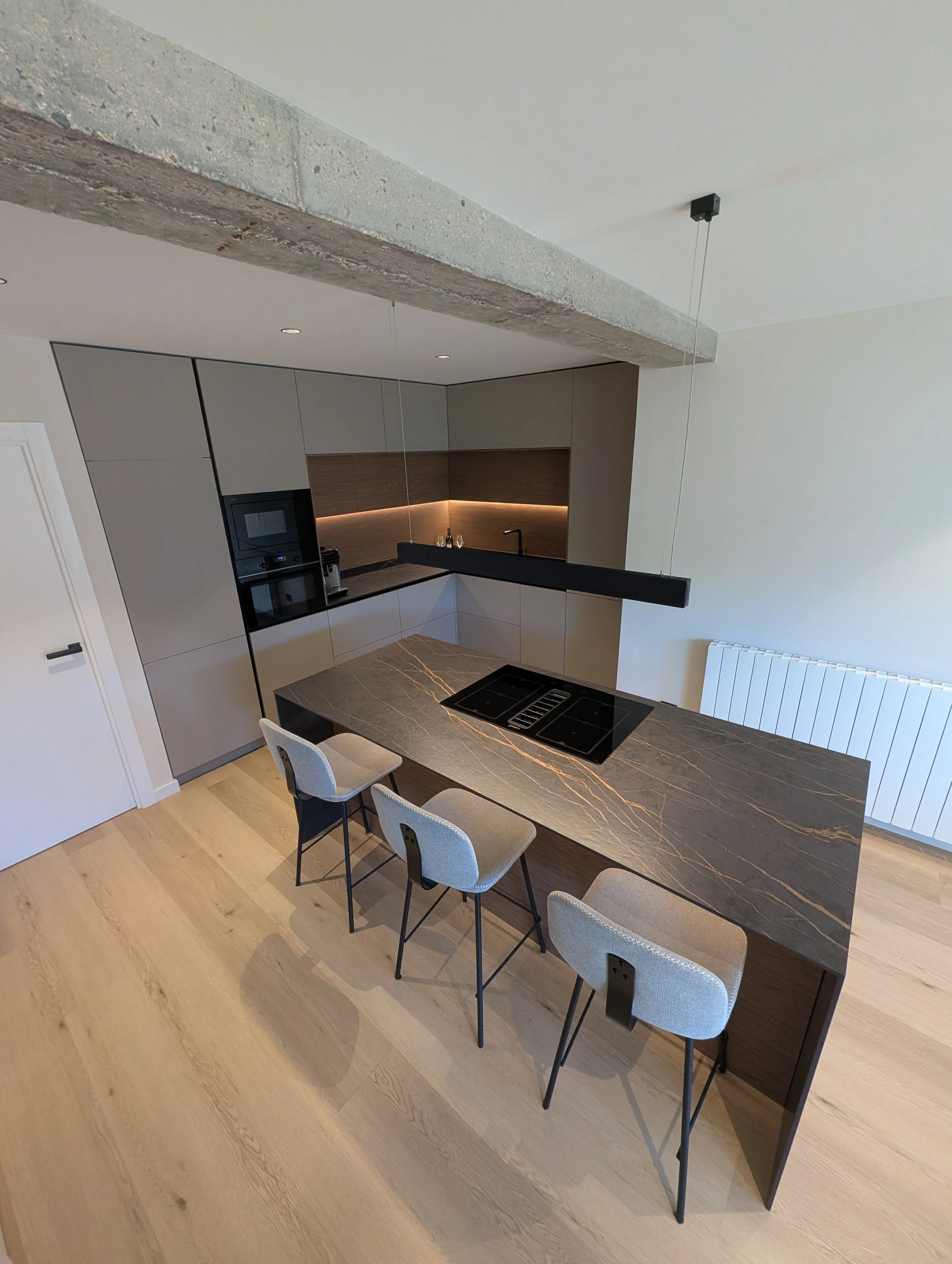 Cocina moderna con encimera de mármol y gabinetes grises, con una barra de desayuno, cuatro sillas de tela gris y una vista parcial de radiador y pared blanca.