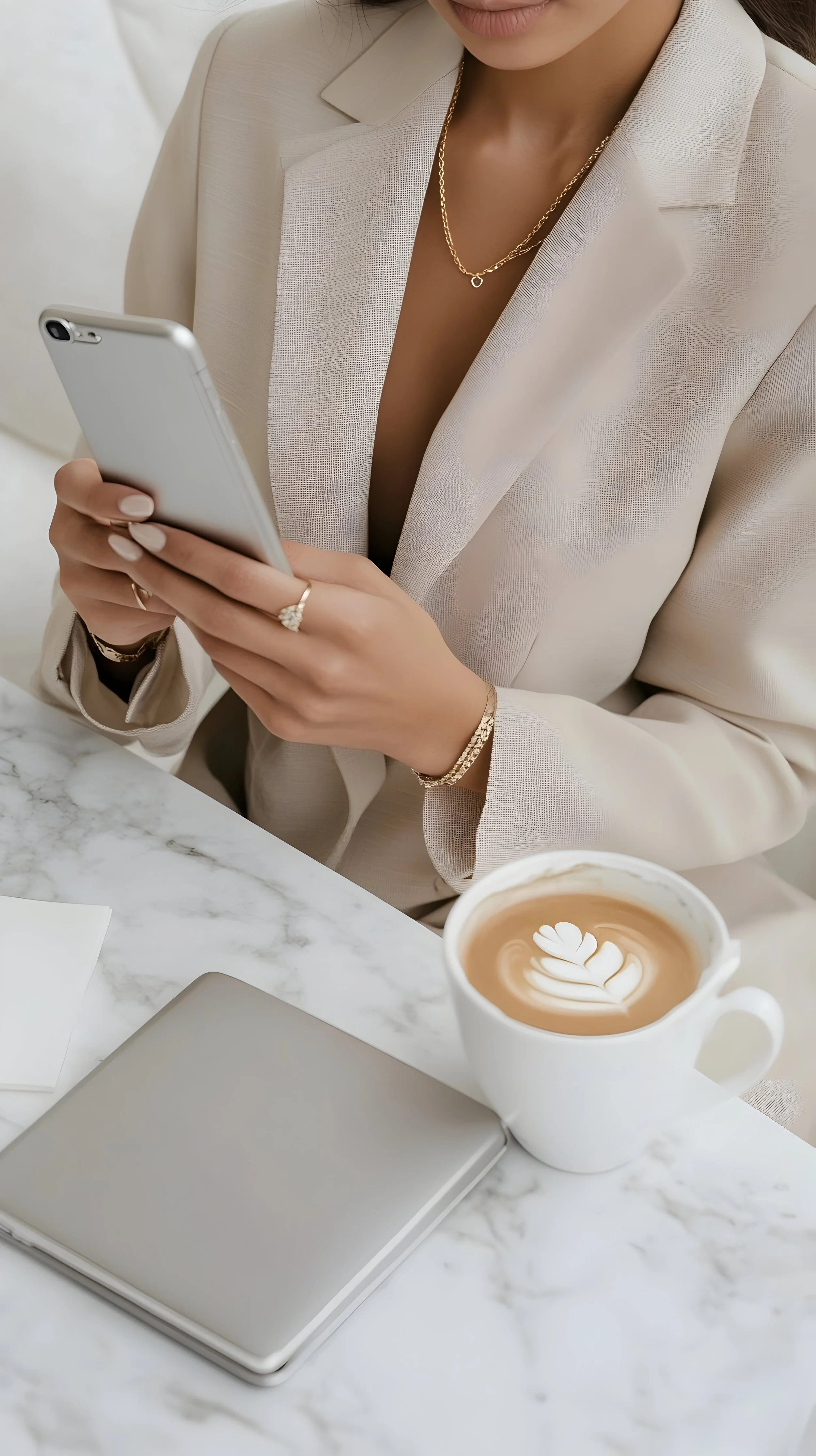Mujer con ropa clara y joyas viste con un blazer, sostiene un teléfono móvil y tiene una taza con un diseño de arte latte en una mesa de mármol interiorista dorleta seoane de vitoria gazteiz y mallorca