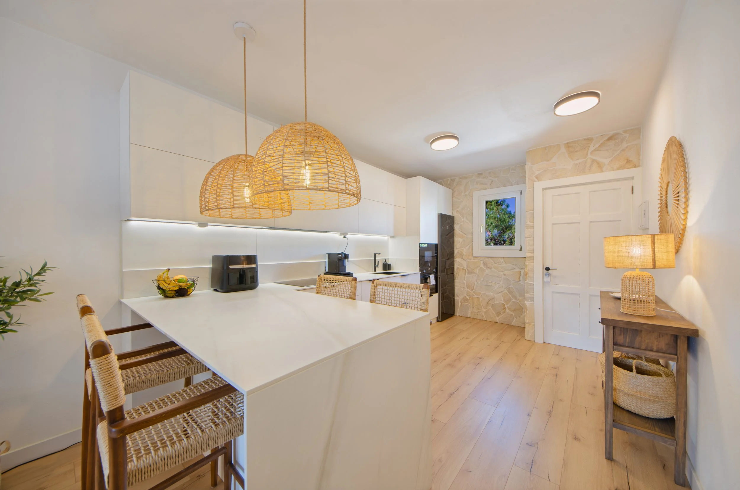 SeoaneinteriorismovitoriagazteizCocina moderna con iluminación cálida, paredes de piedra rosa y muebles blancos, con sillas de mimbre y decoraciones en madera.