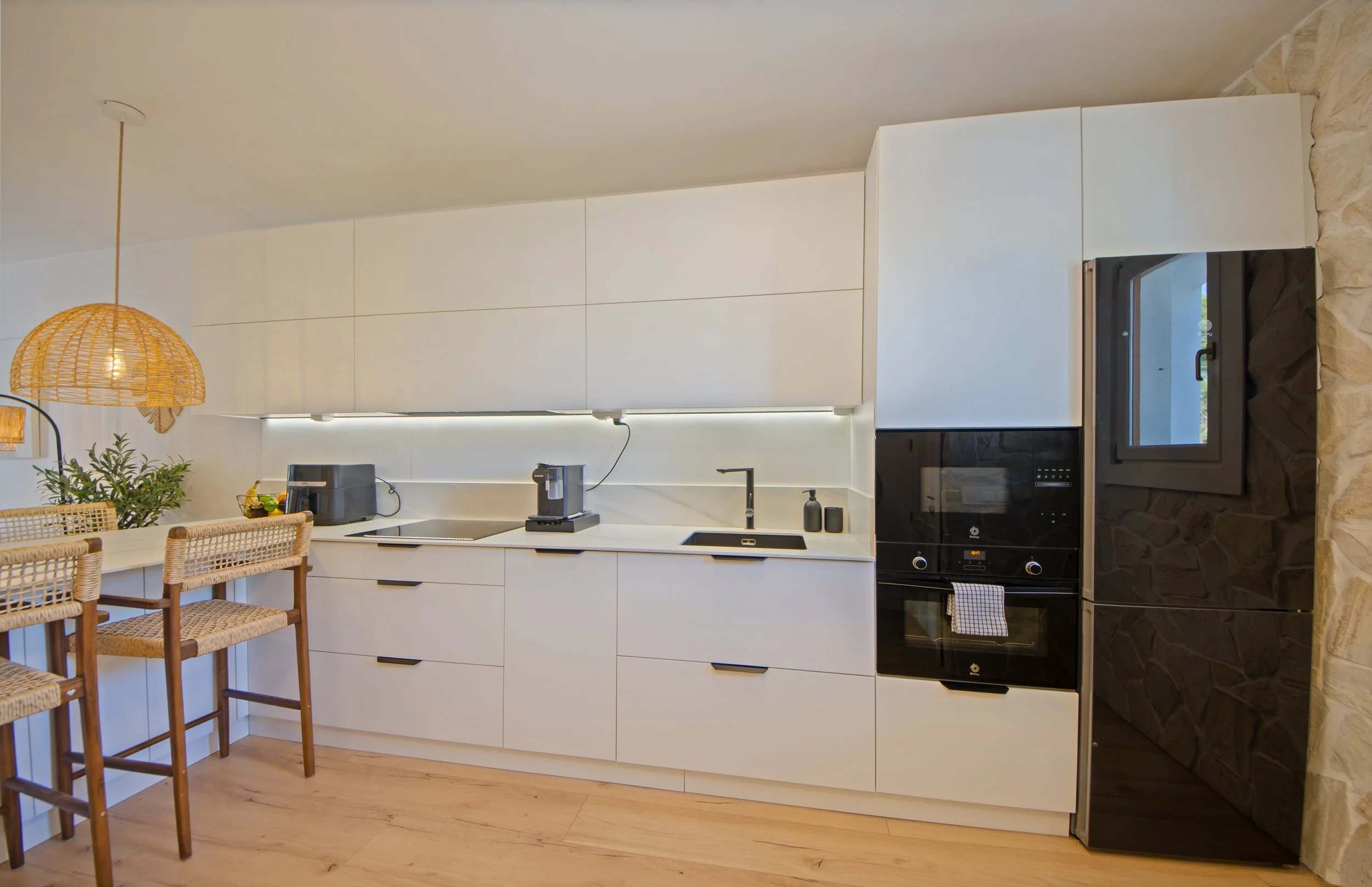 seoaneinteriorismovitoriagazteizymallorcaCocina moderna con gabinetes blancos, horno, microondas y una zona de barra con sillas de mimbre, luminaria de mimbre y una planta en la esquina.
