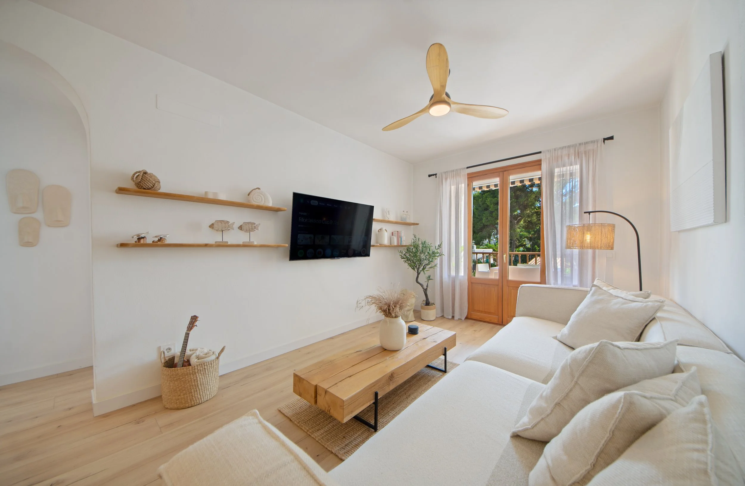 interioristavitoriagazteizSala de estar con paredes blancas, sofá beige y mesa de madera, ventanas con cortinas blancas, estanterías de madera con decoraciones marinas, ventilador de techo y lámpara de pie, con una vista de árboles verdes afuera.