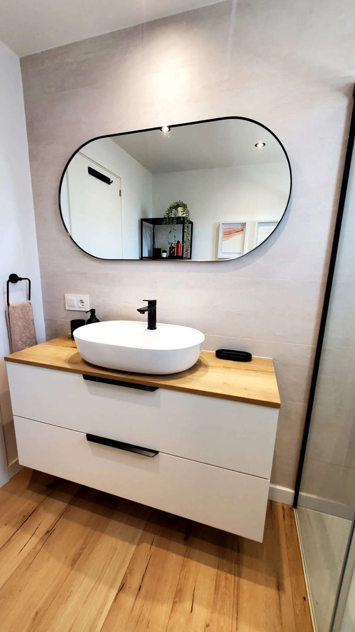 Espejo de forma ovalada sobre un lavabo moderno de cerámica blanca, en un baño con pared gris y piso de madera baño deseñado y fabricado por seoane interiorismo en mallorca. 