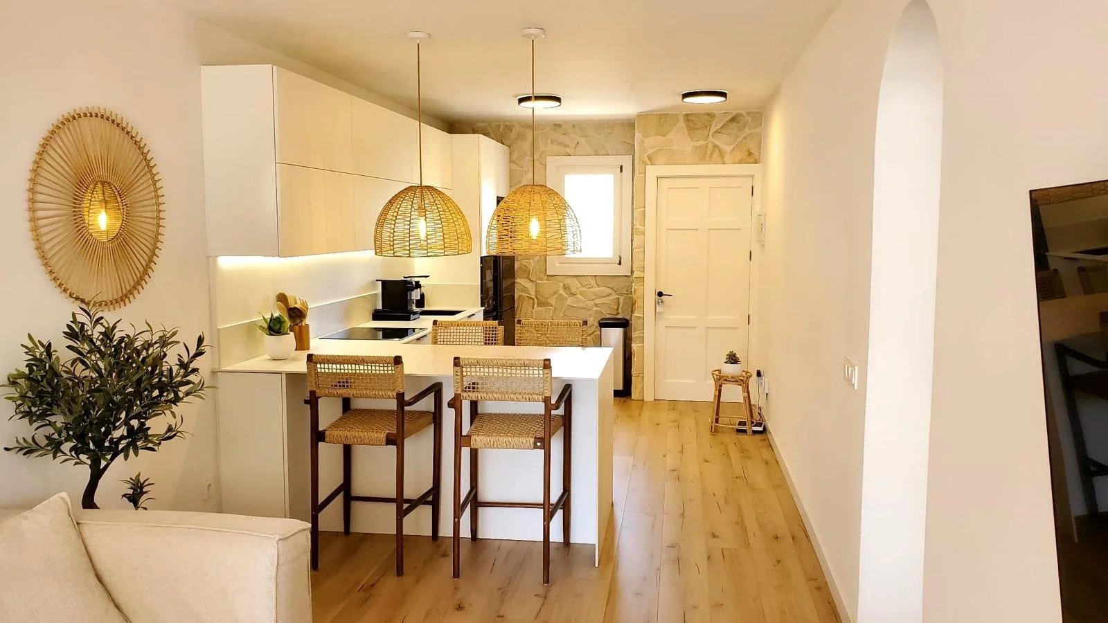 Cocina moderna con encimeras blancas, iluminación empotrada, lámparas de mimbre, paredes de piedra, puerta blanca, estanterías con plantas y utensilios, mesa con sillas de mimbre y madera, suelo de madera diseñada por seoane interiorismo en Mallorca.