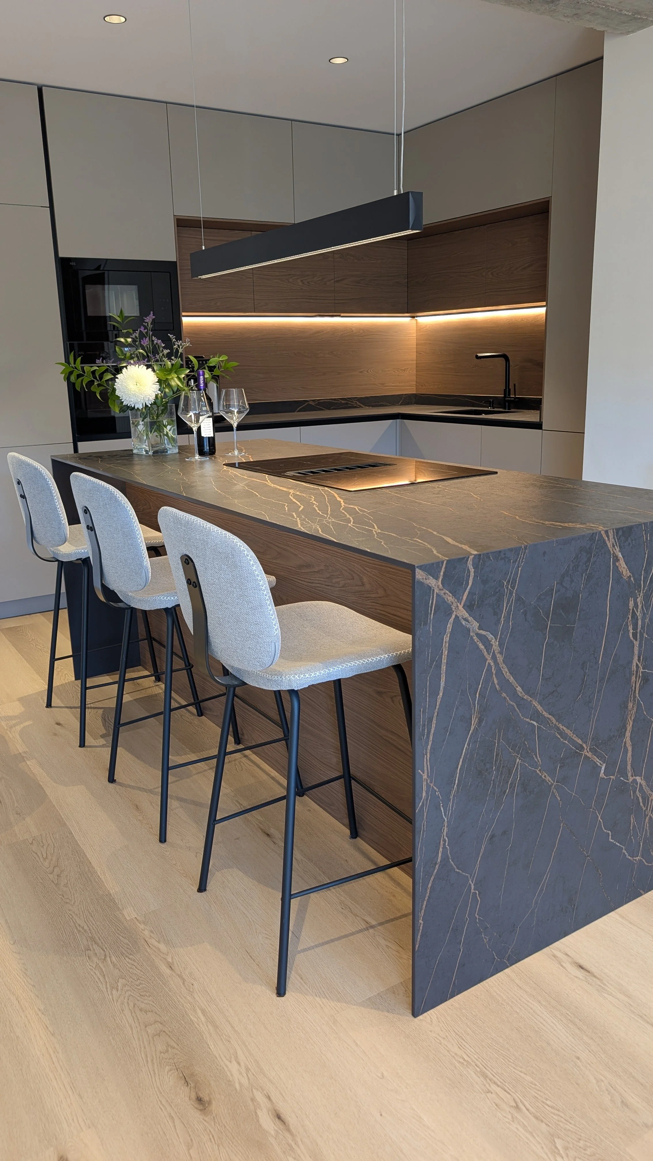 Una cocina moderna con una isla de mármol con vetas doradas, cuatro sillas blancas con patas negras, una especie de jarrón con flores, un vino y dos copas de vino, armarios de madera y gabinete en tono gris claro, iluminación LED en la parte superior