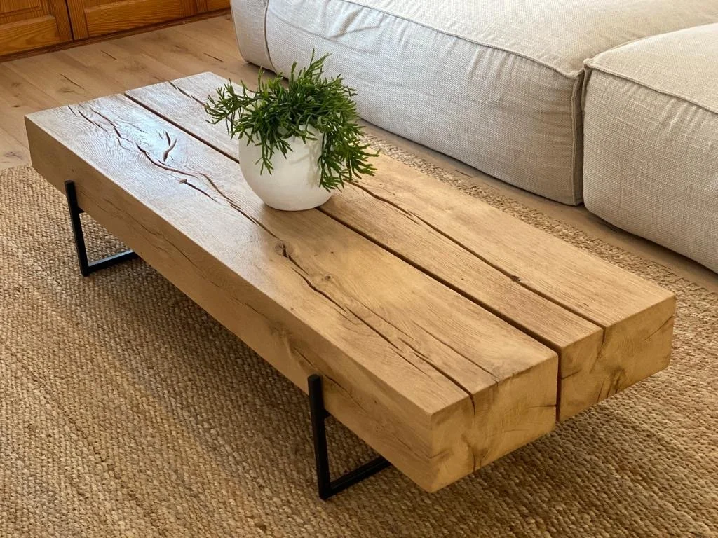 Mesa de madera rústica con detalles de grietas y vetas, sobre patas metálicas negras, en una sala con alfombra y sofá claro.