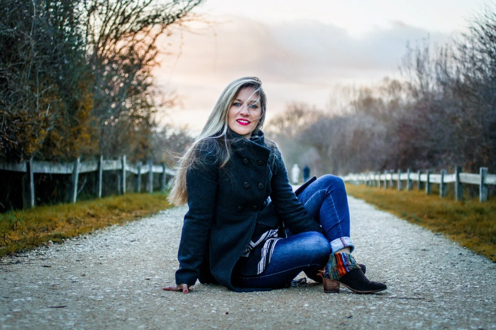 Mujer de cabello rubio sentada en un camino rural con árboles a los lados en un día nublado, vestida con abrigo negro, jeans y botas con calcetas coloridas.