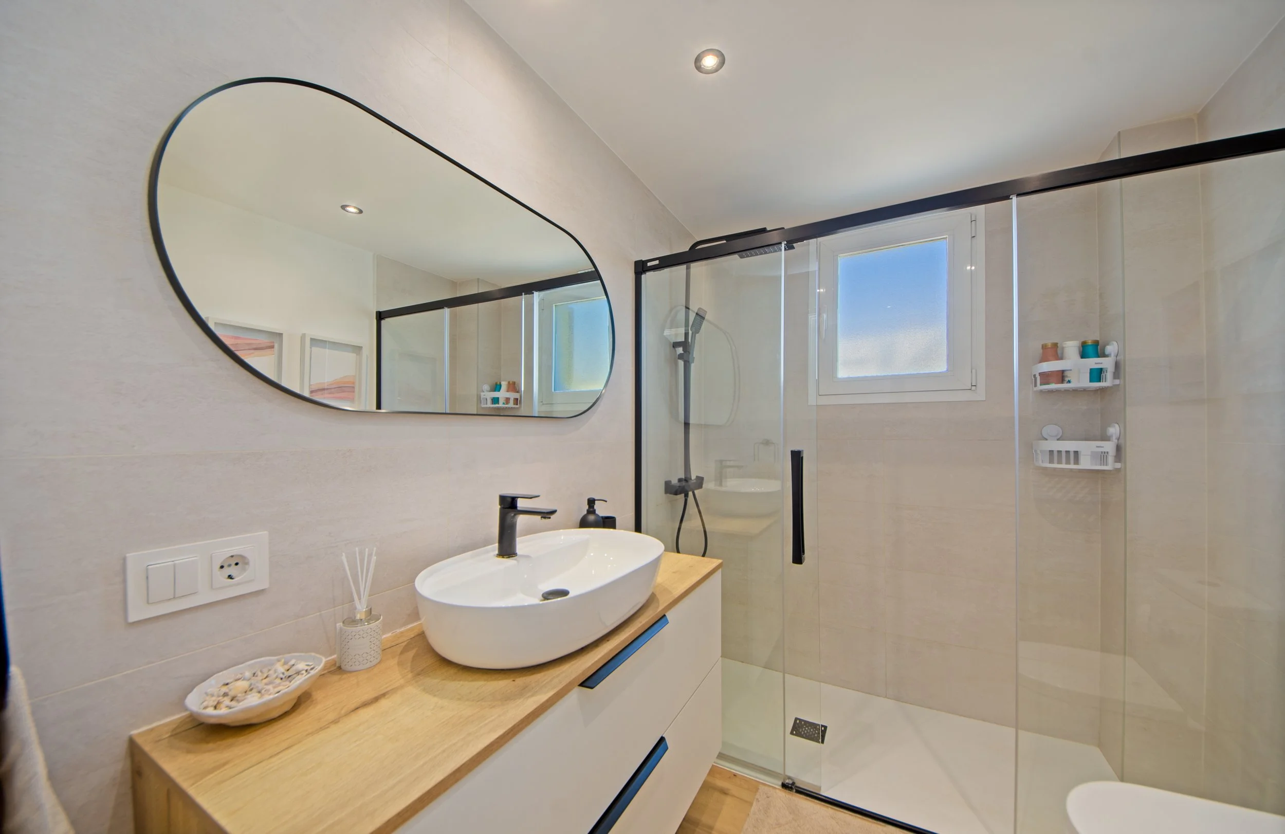 Baño moderno con espejo ovalado, lavabo sobre bancada de madera, ducha con separador de vidrio y ventanas que dejan entrar luz natural.