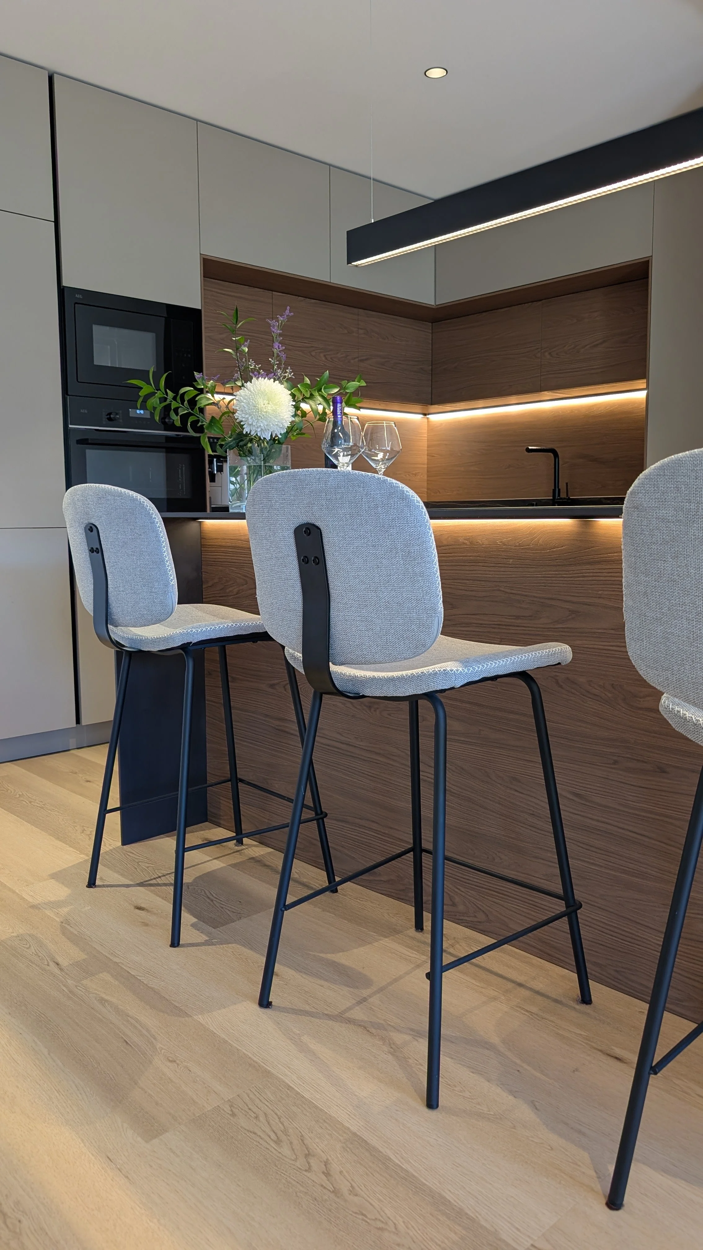 Sala de cocina moderna con barra de madera, flores en una taza de vidrio, dos copas y una botella de vino. Hay sillas con cojines gris claro y estructura de metal negro diseñada por seoane interiorismo en vitoria-gazteiz. 