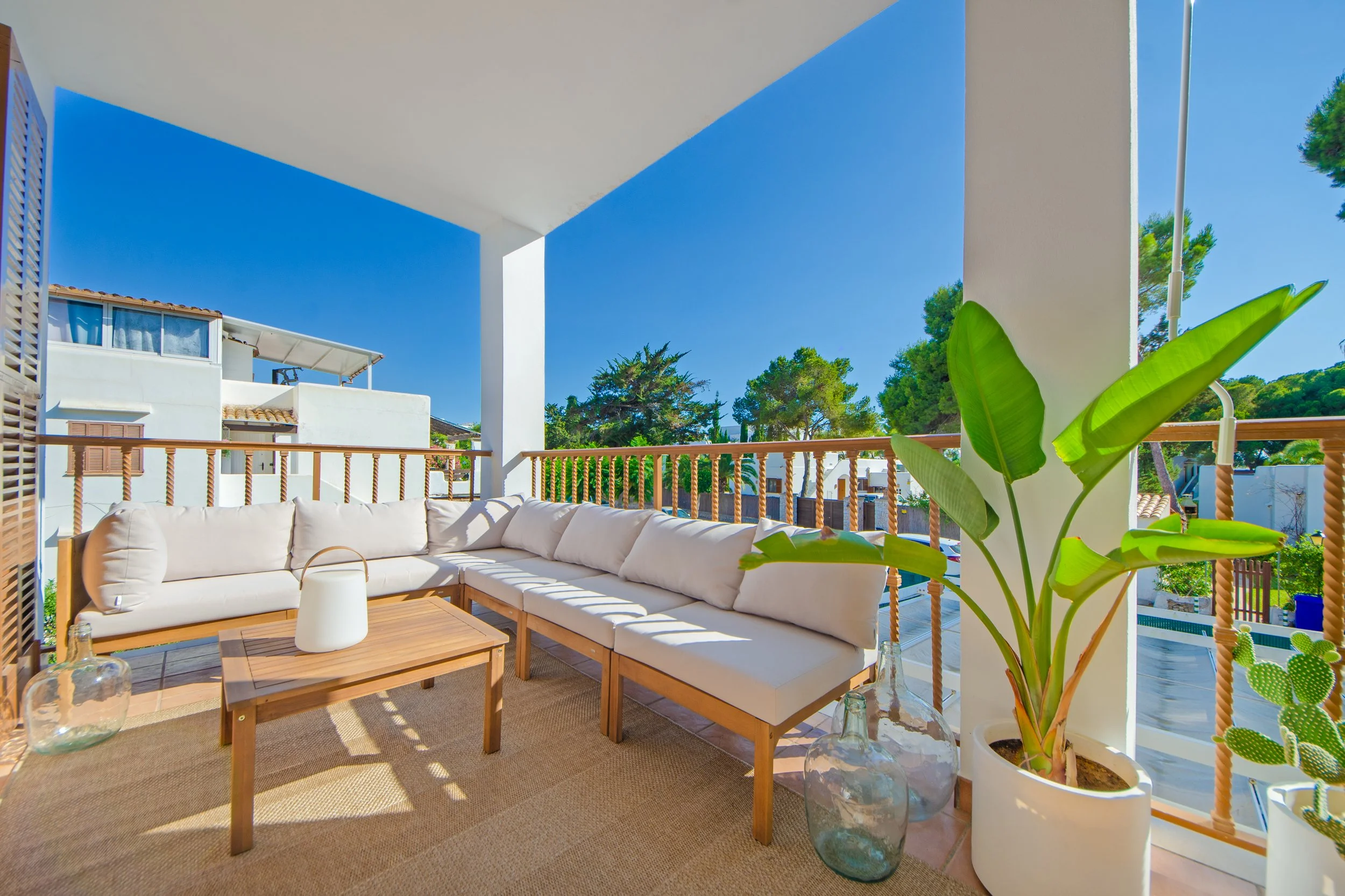 Terraza con sillones blancos en forma de L, mesas de madera, plantas en jarrones de vidrio, barandas de madera, casa moderna con ventanas y azotea, árboles y cielo azul.