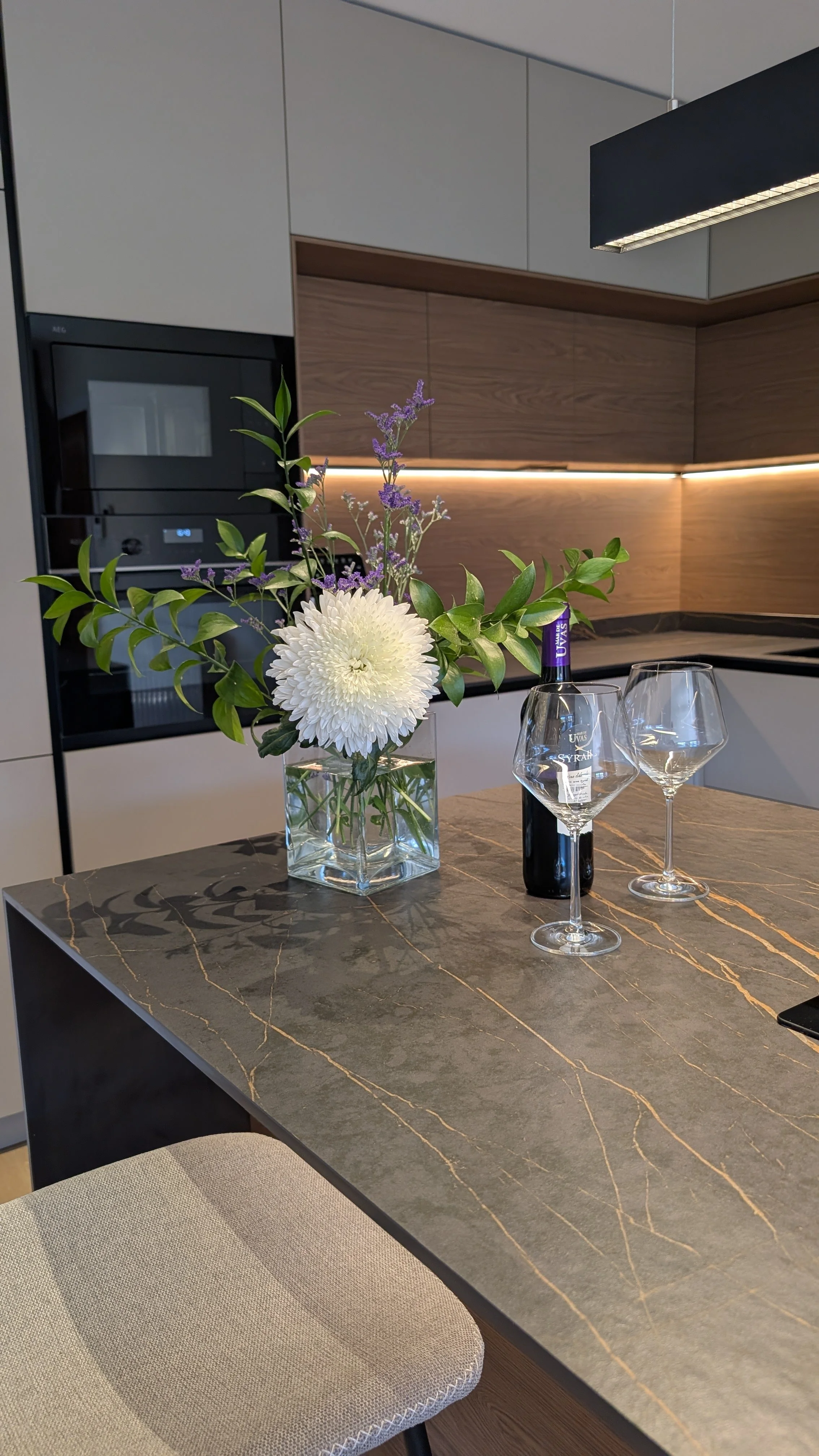 Vaso con flores blancas y verdes, botella de vino y dos copas en una encimera de cocina de granito oscuro con líneas doradas, en un ambiente moderno con gabinetes de madera y paredes blancas.