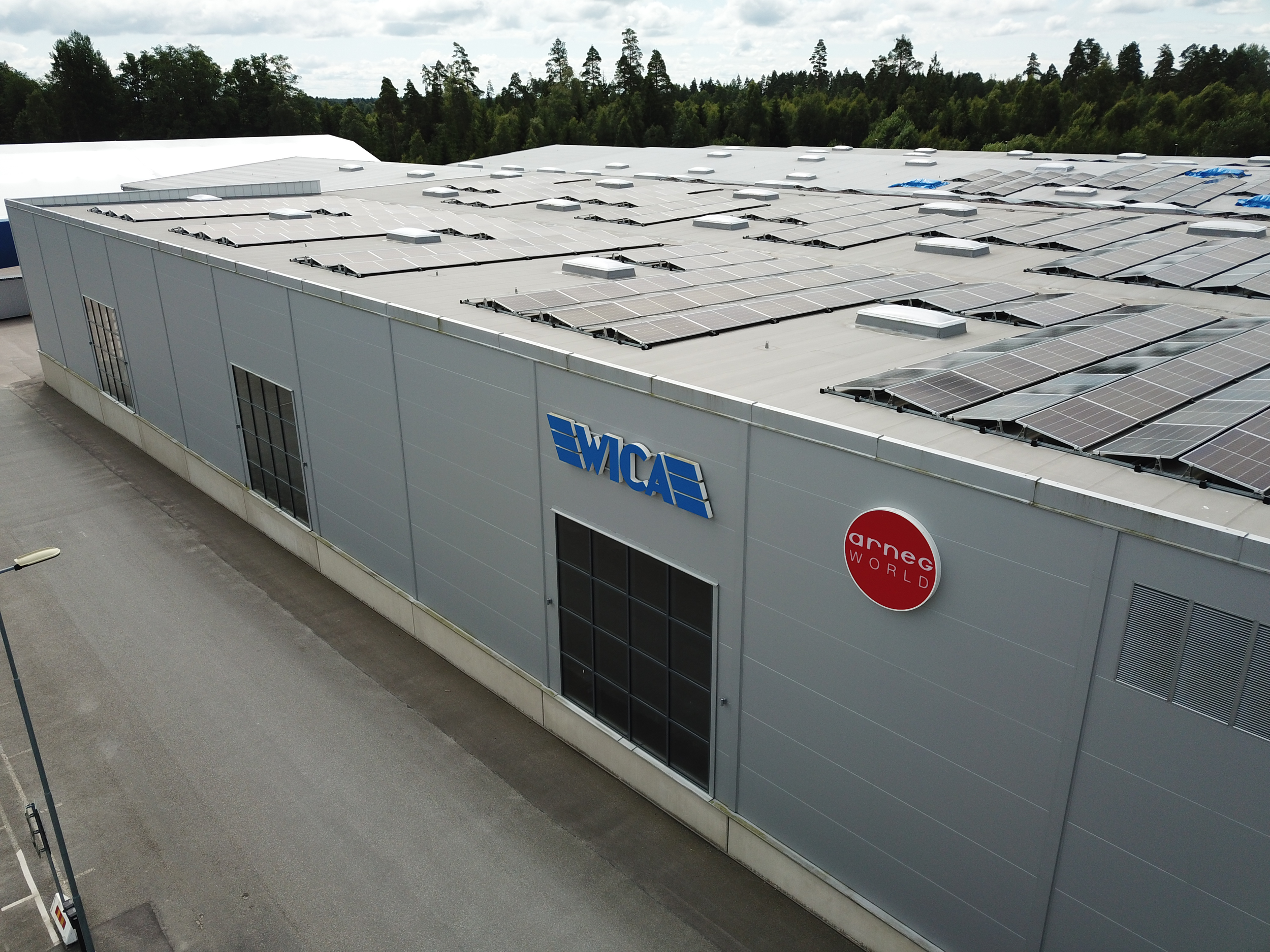 Suuri teollisuusrakennus, jonka seinässä on logot WMCA ja Arne G World, katolla on aurinkopaneeleita, ja se sijaitsee teollisuusalueella metsän ja kävelytien vieressä.