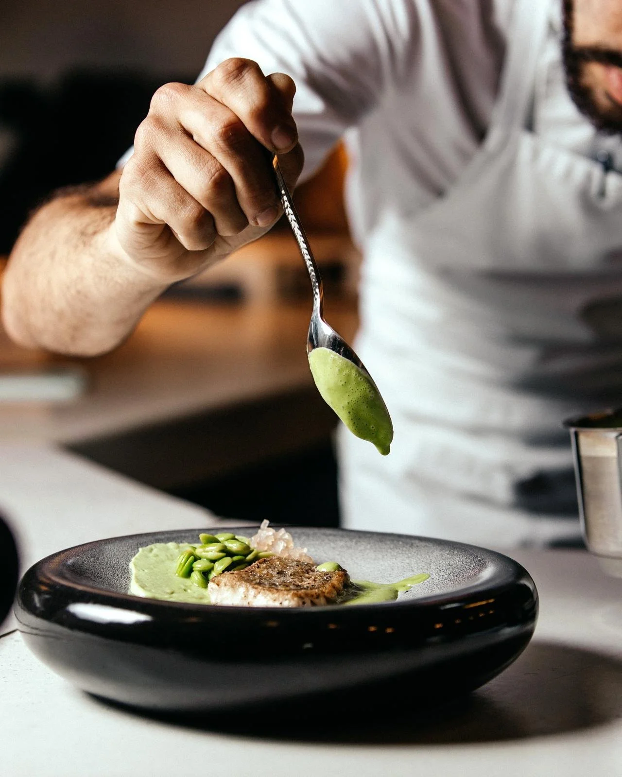Chef garnishing a plated dish with green sauce and vegetables in a modern kitchen.