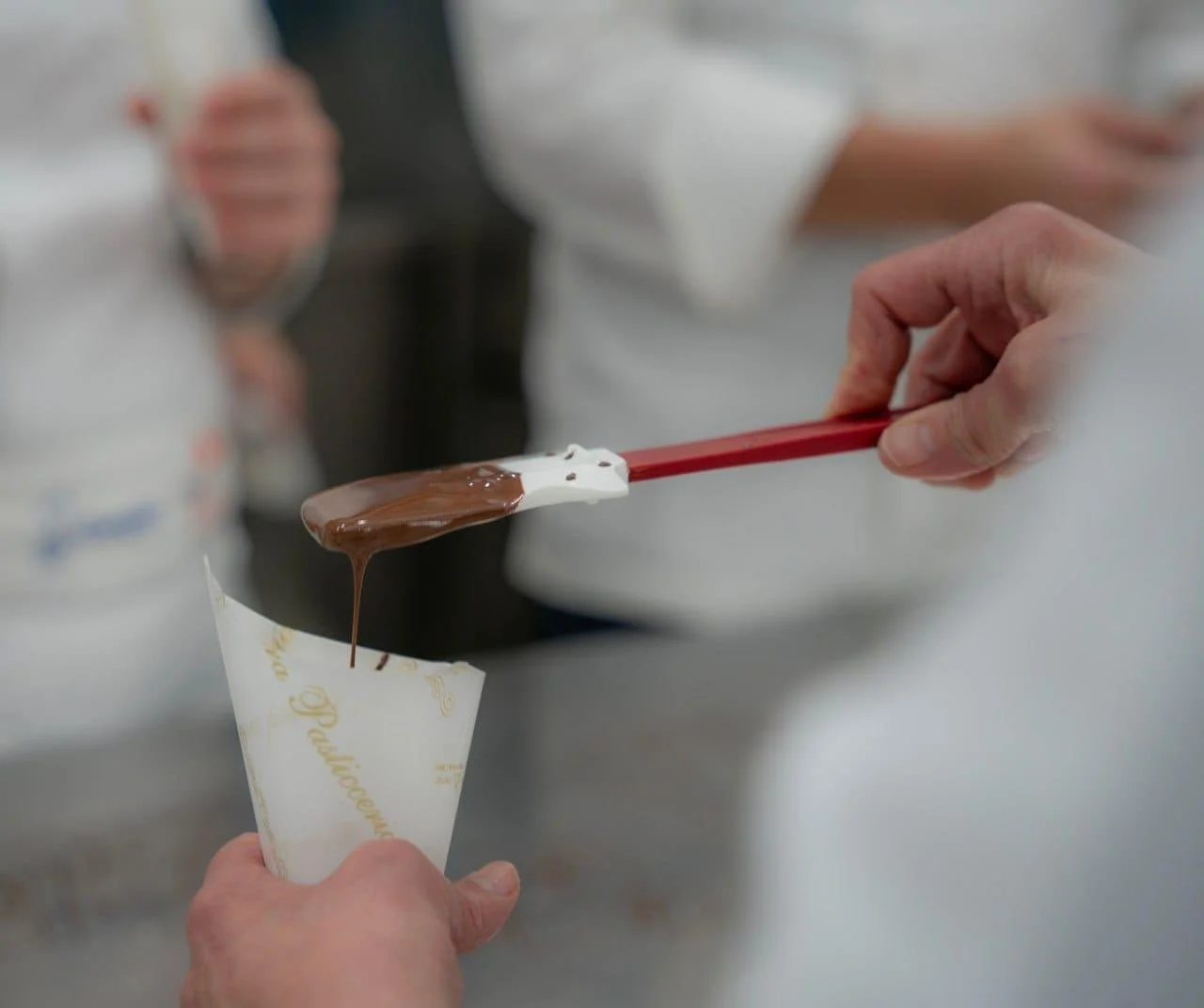 A person using a spatula to fill a cone with melted chocolate, in a busy kitchen environment.