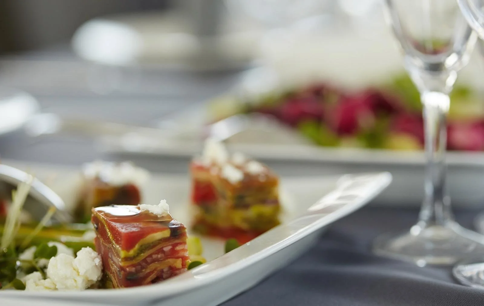 Close-up of layered terrine with meat and vegetables on a white platter, garnished with herbs and cheese, with a blurred floral arrangement in the background.