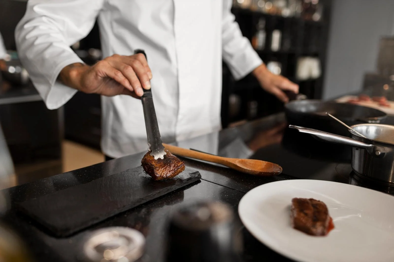 Chef in a white coat using tongs to serve a piece of cooked meat on a black slate plate in a professional kitchen.
