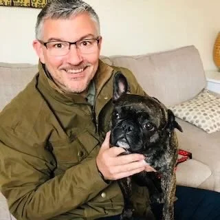 A man with gray hair and glasses smiling, holding a black and brindle French Bulldog inside a living room with a beige sofa and patterned pillows.