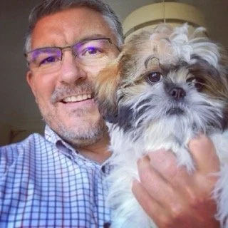A man with glasses smiling and holding a small fluffy dog.