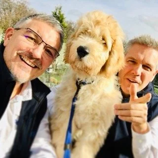Two smiling men and a large, fluffy dog outdoors with trees and sky in the background.