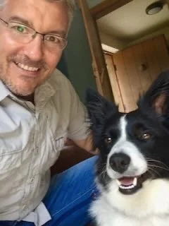 A man smiling and posing with a black and white dog indoors near a wooden door.