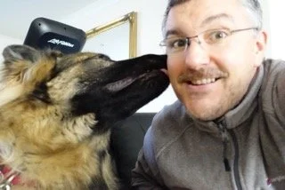 A man smiling while a dog sniffs his face.