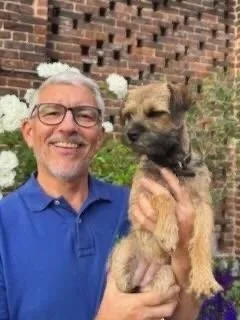 A man with gray hair, glasses, and a blue shirt smiling while holding a small brown and black puppy in front of a brick wall with white flowers.