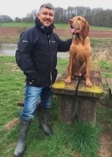 Man in black jacket and jeans standing next to a large brown dog on a wooden platform outdoors.