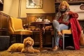 A woman with curly hair and a dog sitting on the floor in a cozy, vintage-style living room with a fireplace, mirror, and yellow armchair.