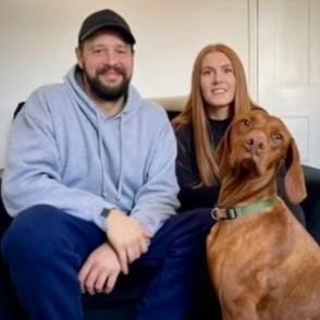 A man with a beard wearing a baseball cap and a woman with long hair sitting on a couch with their brown dog.