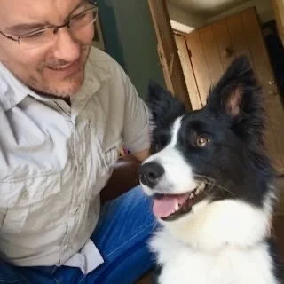 A man smiling with a happy black and white dog inside a home.