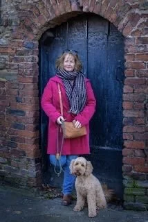 A woman in a pink coat standing in front of a dark blue wooden door with a small beige dog on a leash.
