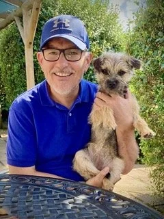 A man wearing a blue cap, glasses, and a blue polo shirt smiling while holding a small, scruffy dog outdoors.