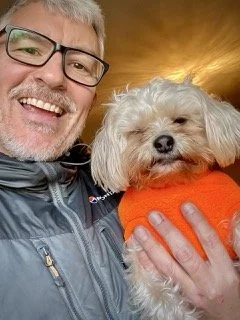 A man smiling while holding a small, fluffy dog wearing an orange sweater, against a warm, golden sunset background.