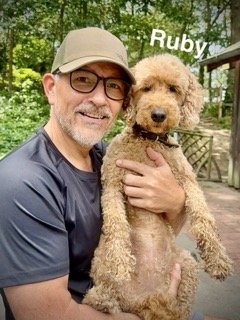 Man wearing glasses and a beige cap holding a curly-haired dog outdoors in a wooded area