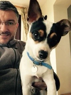 A man holding a small black and white puppy with large ears and expressive eyes indoors.