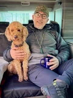 A man with glasses and a beard sitting on a bus seat, holding a small curly-haired dog and a mug.