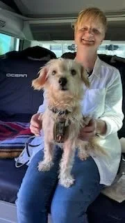 A smiling woman sitting inside a vehicle holding a small scruffy dog with light-colored fur.