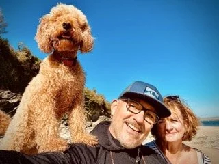 A man and woman taking a selfie outdoors with a fluffy dog on a sunny day at the beach.