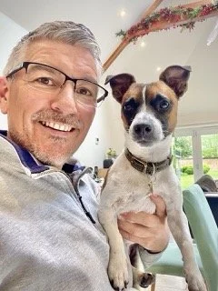 A man with glasses and a beard smiling, holding a small dog with a brown and white coat and pointy ears, inside a bright room with large windows and a green chair in the background.