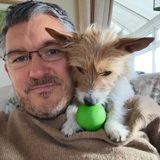 Man holding a small dog with a green ball in its mouth, sitting on a couch near a window.