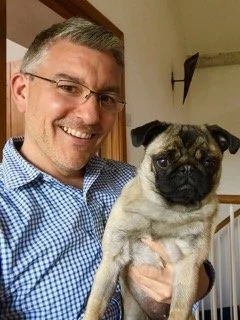 A man smiling while holding a small pug dog indoors.