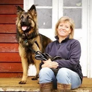 A woman sitting on the ground next to a large Belgian Malinois dog outside, holding a leash, with a red wooden building behind them.